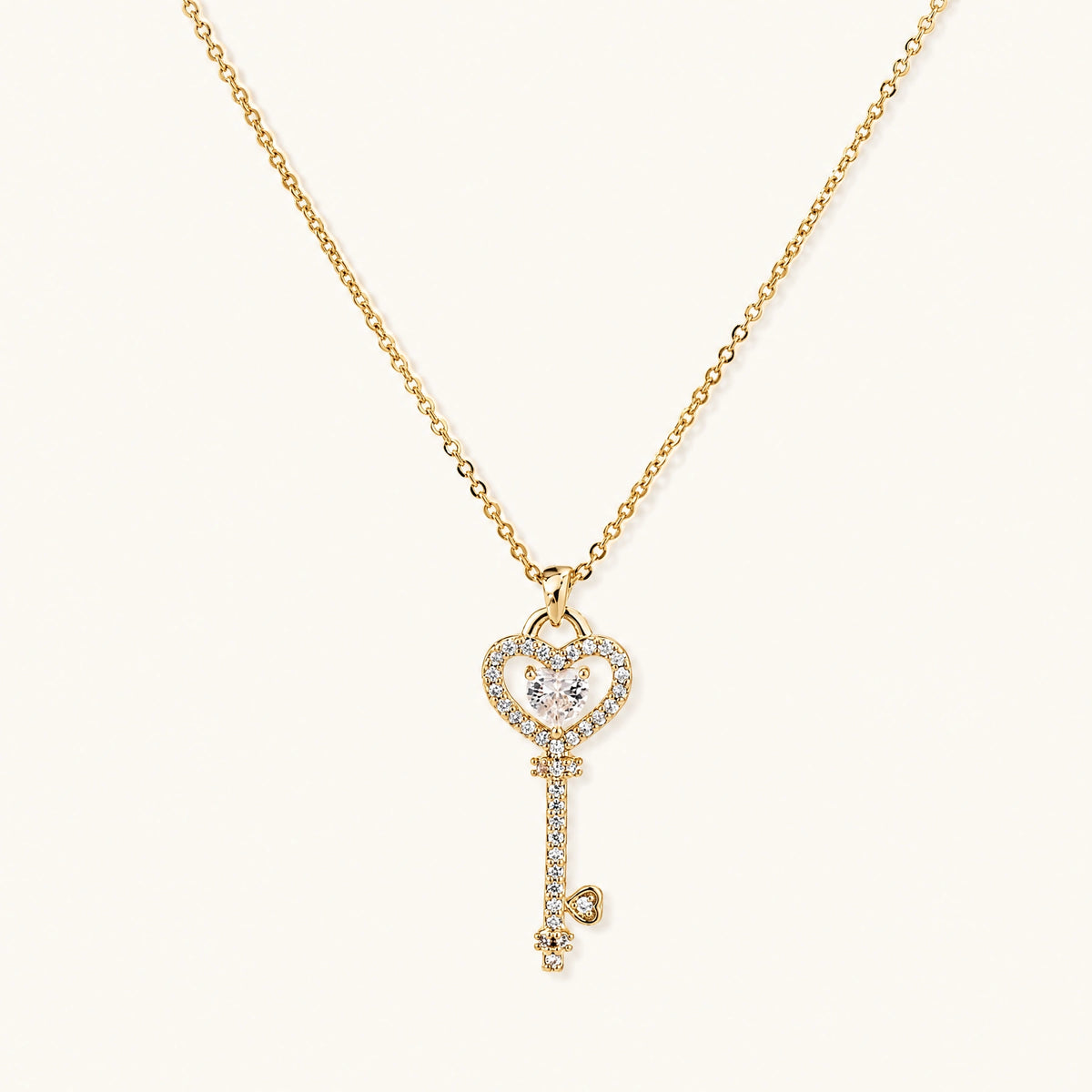 Delicate gold key pendant with heart-shaped bow and crystal pavé detailing on fine chain against cream background.