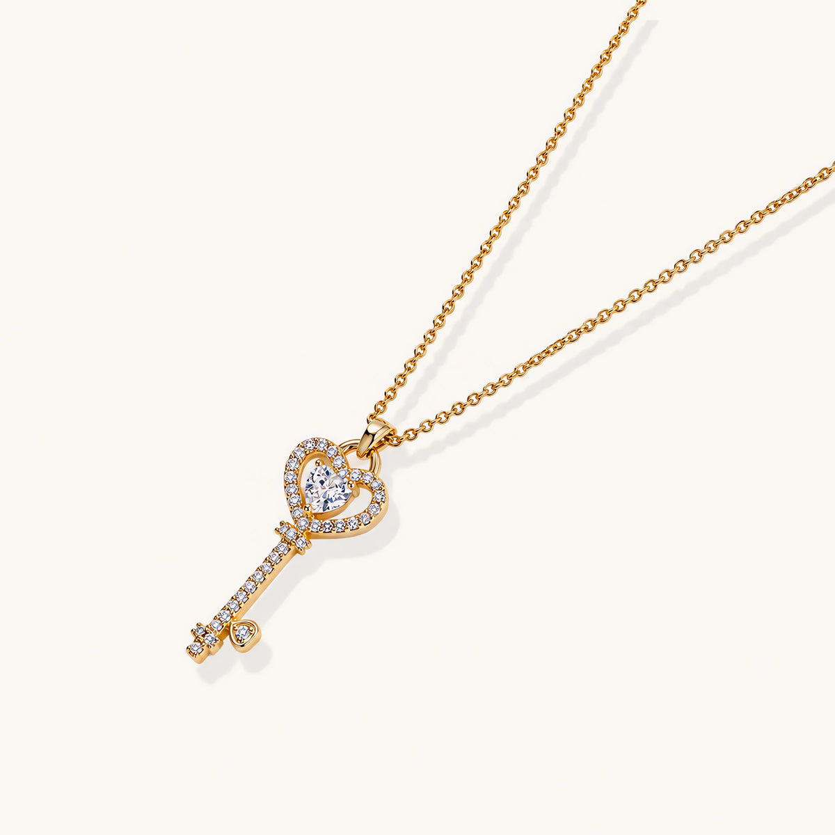 Delicate gold key pendant with heart-shaped bow and crystal pavé detailing on fine chain against cream background.