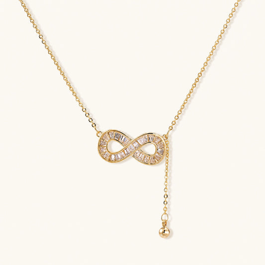Gold infinity pendant with baguette crystals on adjustable chain against cream background.