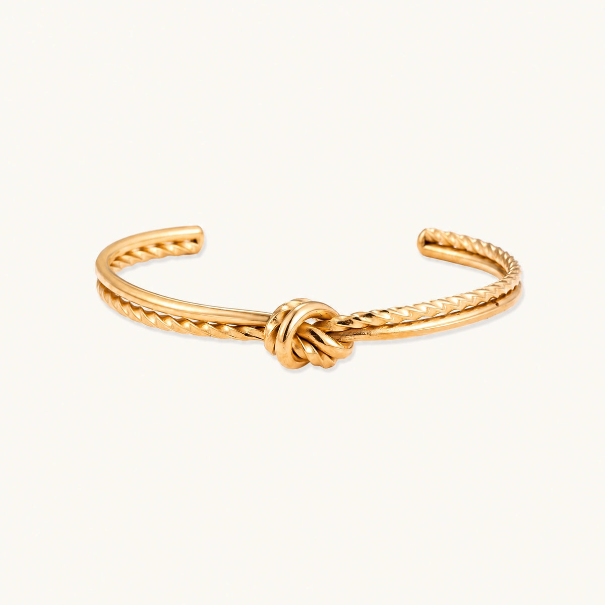 Gold cuff bracelet with twisted rope texture and decorative knot centerpiece on cream background.