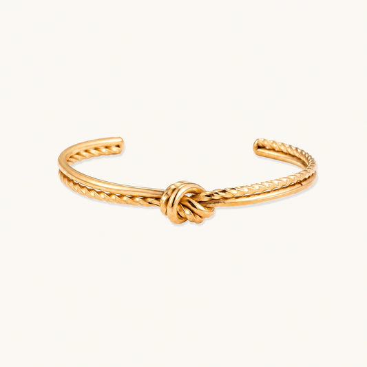 Gold cuff bracelet with twisted rope texture and decorative knot centerpiece on cream background.